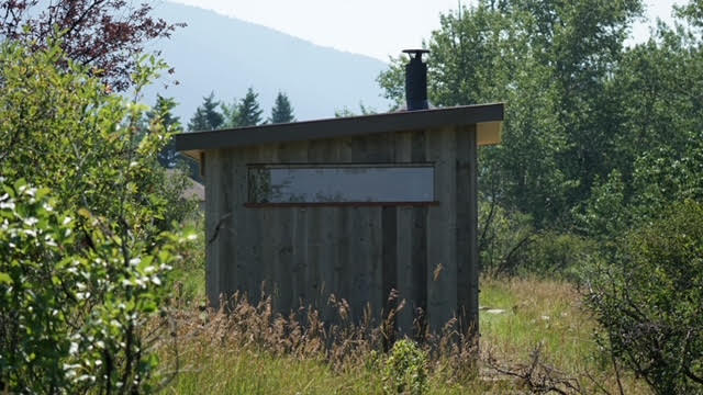 Grey corrugated-metal sauna with a stovepipe chimney in an open grassy field, mountains in the distance